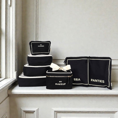 Black Bag-all Travel Glam 4-Pack with labeled makeup, charger, bra, and panties organizers on white ledge near window