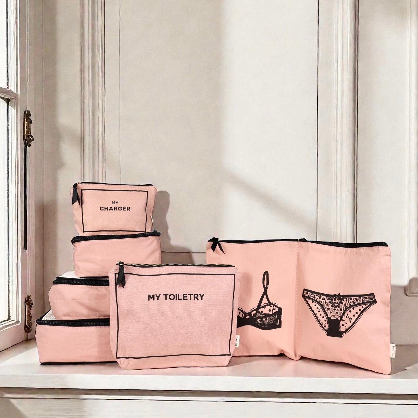Pink Bag-all Weekend Essential Packing Set 4-Pack with labeled cotton cubes for toiletries, chargers, and lingerie on window ledge