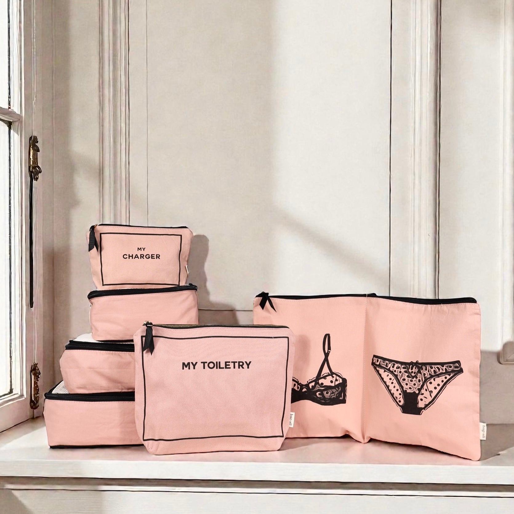 Pink Bag-all Weekend Essential Packing Set 4-Pack with labeled cotton cubes for toiletries, chargers, and lingerie on window ledge