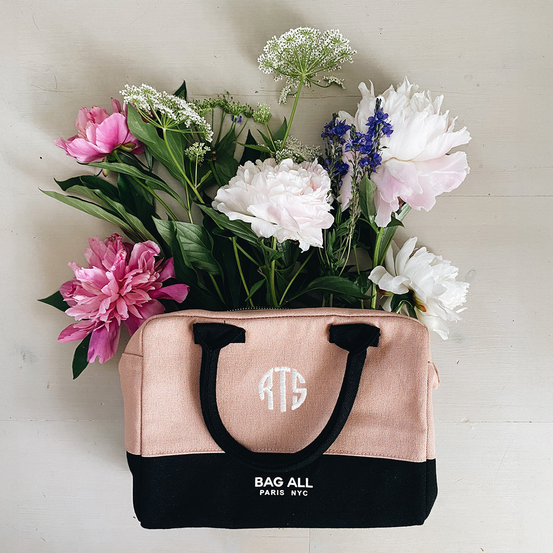 Bag-all Stylish Monogram Lunch Box in blush pink and black with monogram detail, filled with fresh spring flowers including peonies, delphiniums and Queen Anne's lace against a light background