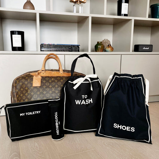 Bag-all Toiletry Travel Gift Set 4-Pack in black cotton featuring toiletry pouch, toothbrush case, shoe bag and wash bag, displayed with designer luggage on modern shelf