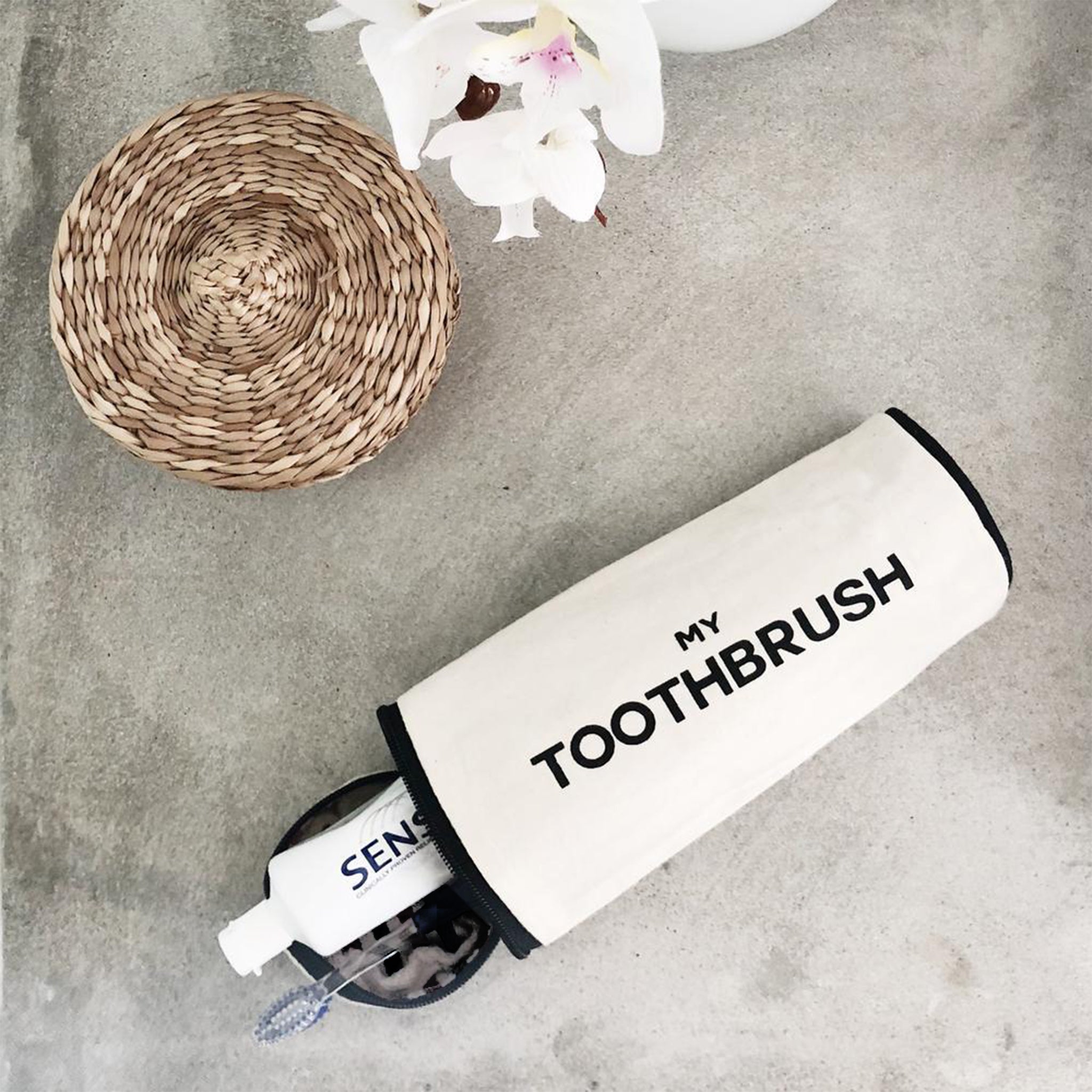 Bag-all My Toothbrush Case in cream cotton, part of Toiletry Travel Gift Set Deal 4-Pack, shown with decorative woven coaster and orchid flower on gray surface