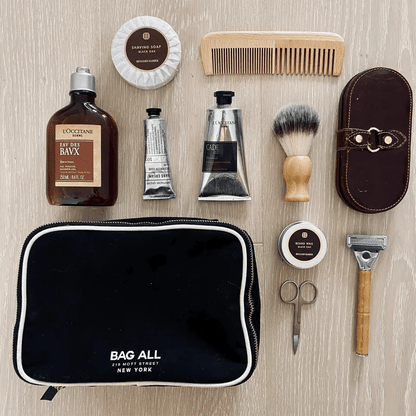 Bag-all Double Sided Toiletry Case in Black shown with grooming essentials including shaving brush, comb, lotions, and travel-sized toiletries on wooden surface, featuring waterproof lining and dual zipper design