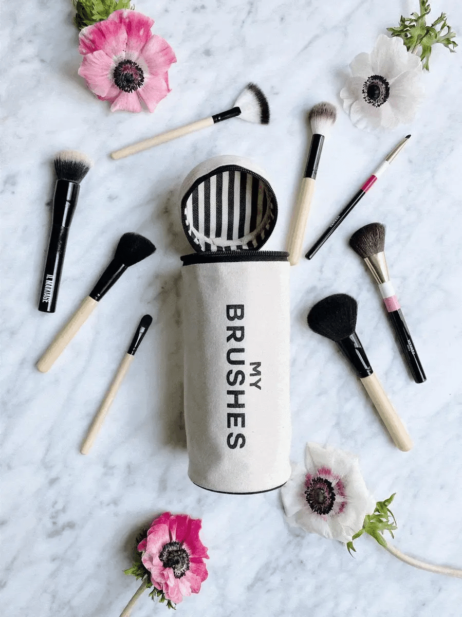 Bag-all Brushes Cylinder Case in cream with striped interior, displayed with makeup brushes and anemone flowers on marble surface, showcasing stylish organization solution