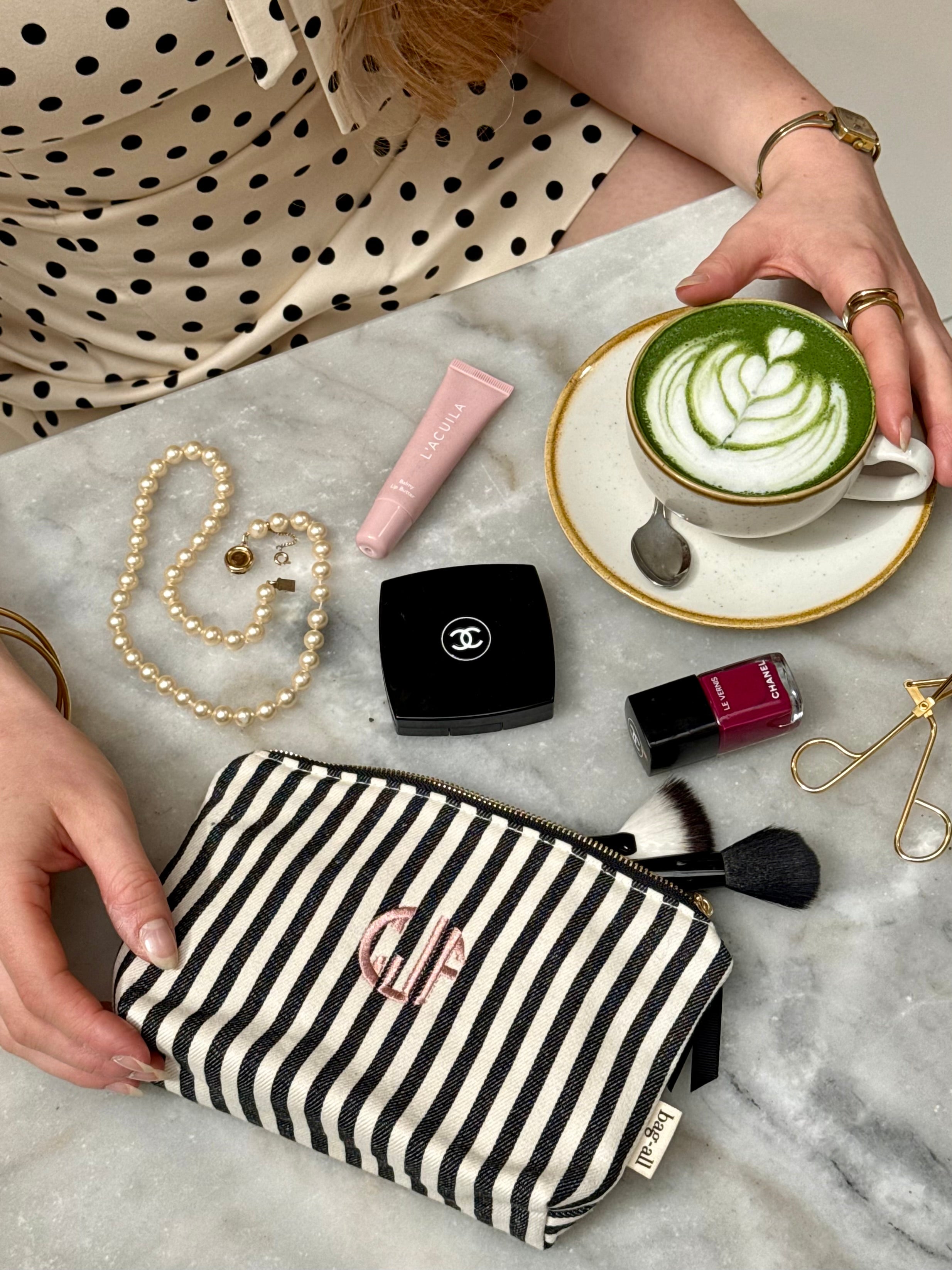 Woman holding striped cotton pouch with pink monogram next to makeup, pearl necklace, and cup of matcha on marble table by Bag-all