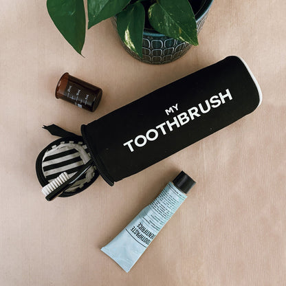 Bag-all black cylindrical toothbrush case with white text, zipper open showing striped interior, styled with tubes and plant nearby