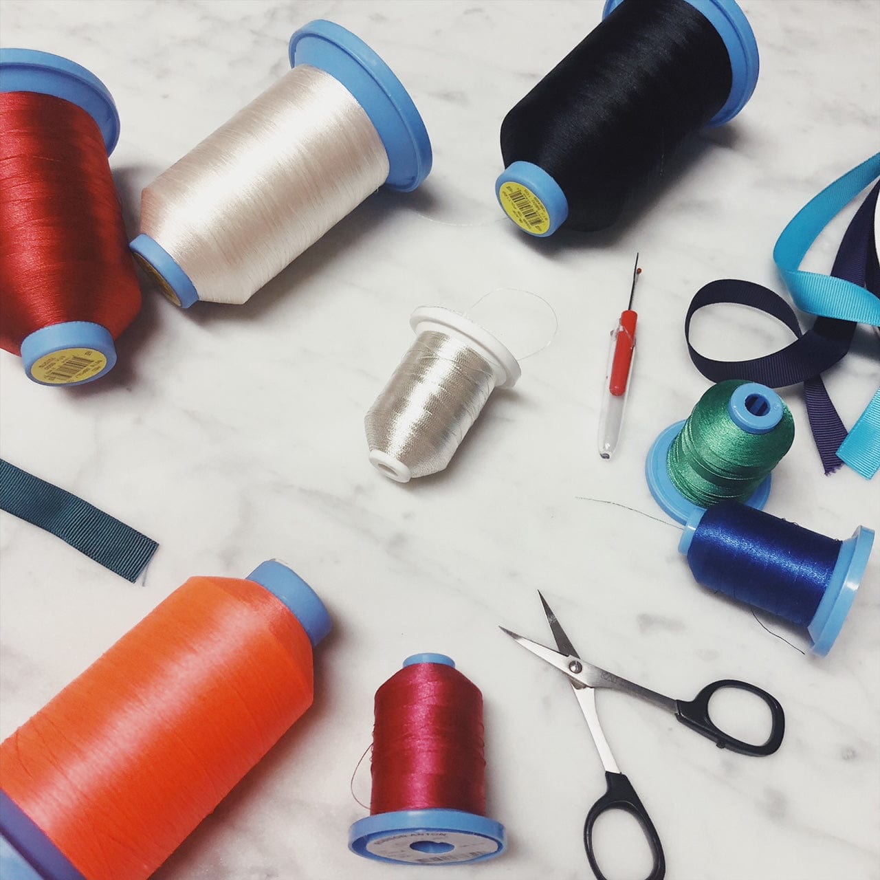 Bag-all monogramming supplies featuring colorful thread spools in red, black, white, orange, and blue, with scissors and ribbon for personalized bag customization service