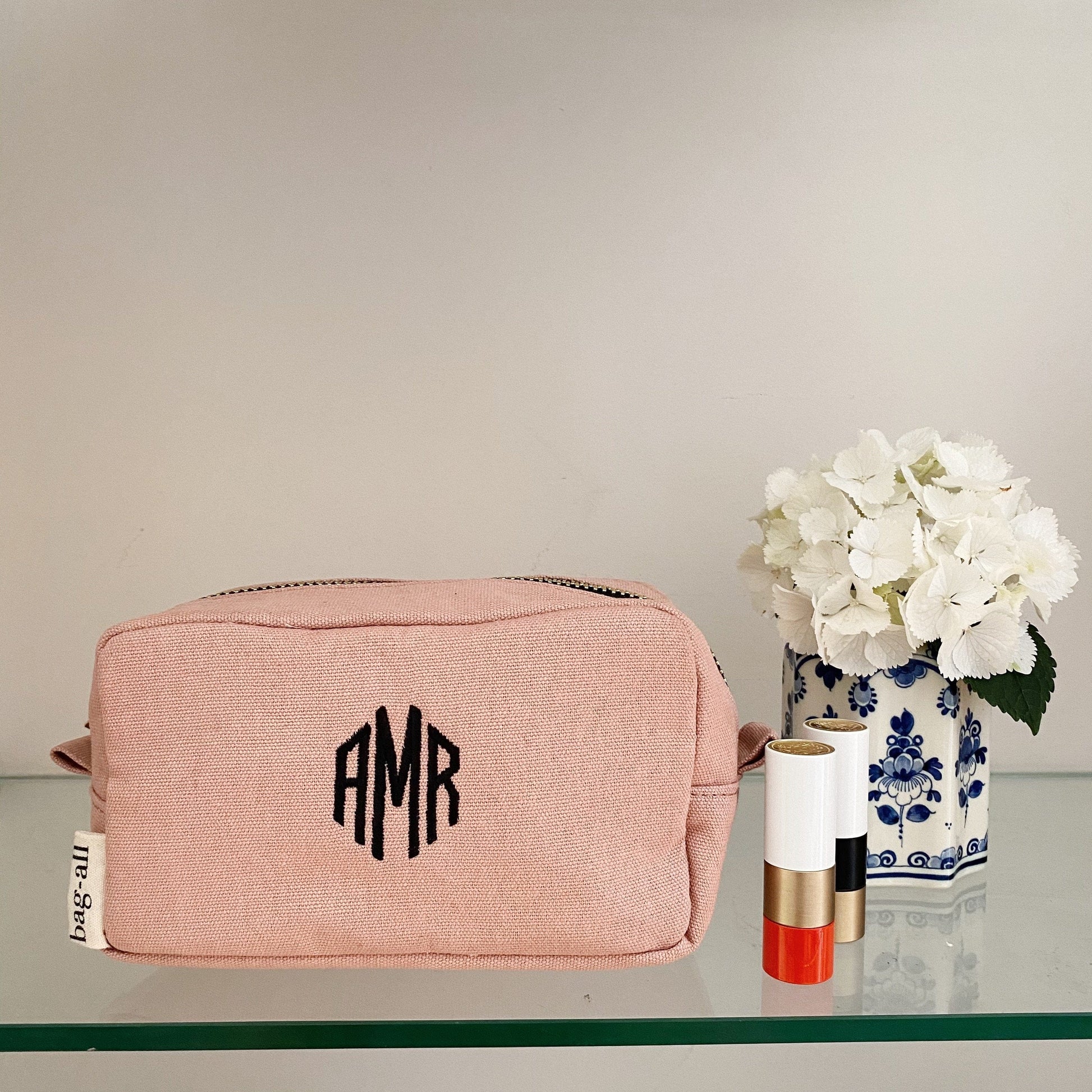 Bag-all personalized pink cotton cosmetic bag with black monogram initials, displayed next to white hydrangeas in blue and white vase and lipstick on glass surface