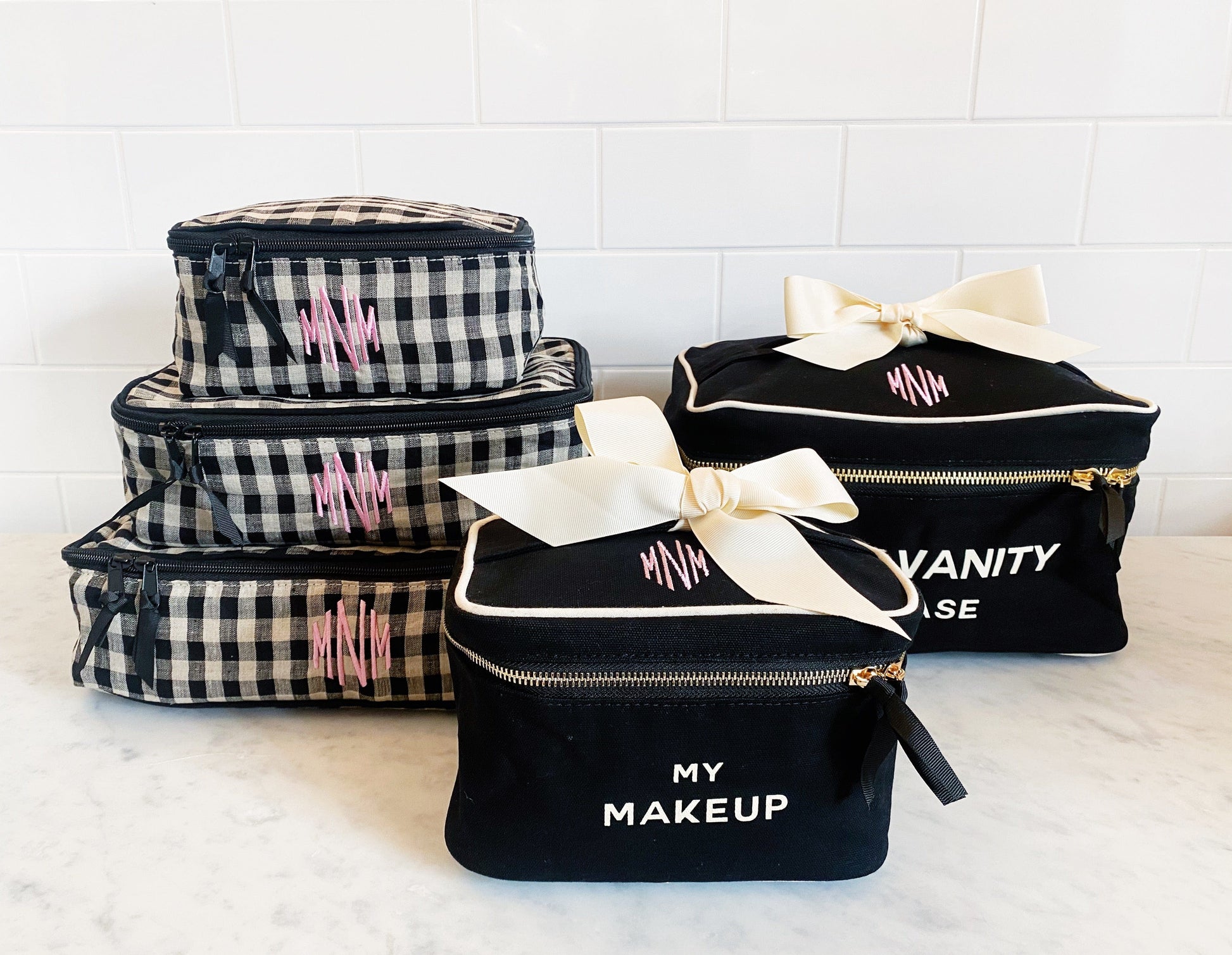 Bag-all personalized monogram travel cases in black gingham and solid black, featuring makeup and vanity cases with customizable initials and elegant bow details
