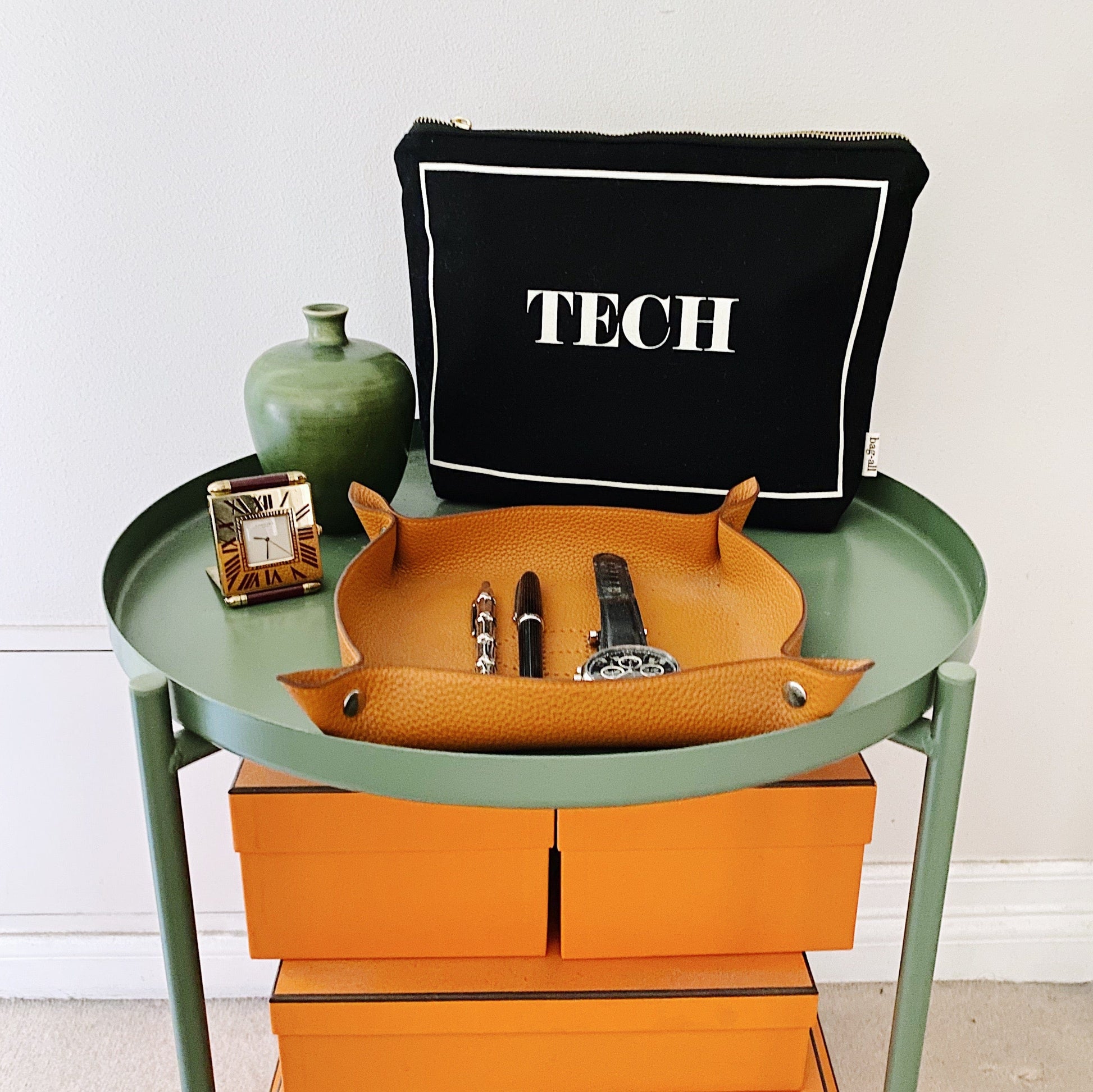 Bag-all Black Tech Pouch organizer displayed on green side table with tan leather catchall tray, showcasing stylish electronic accessory storage solution