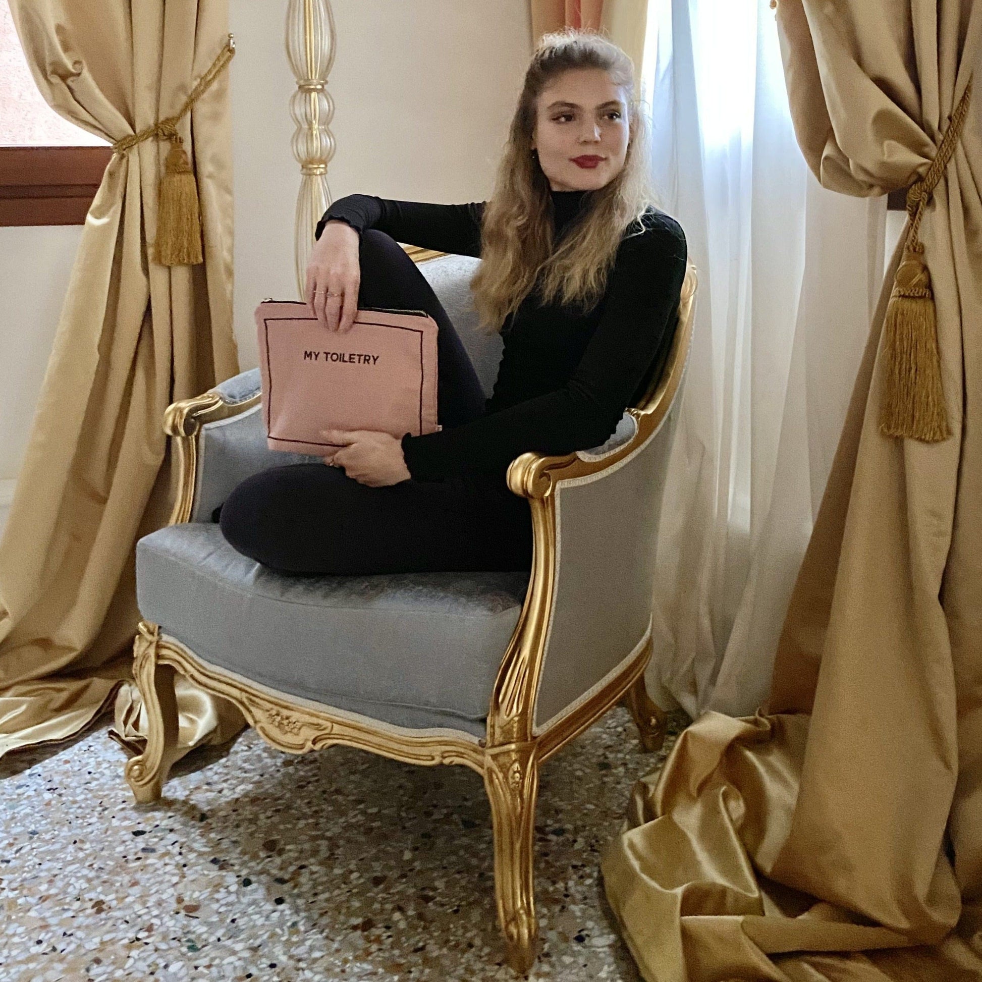 Bag-all Toiletry Pouch with Coated Lining in blush pink shown on elegant vintage chair, featuring waterproof interior and metal zipper for stylish travel organization