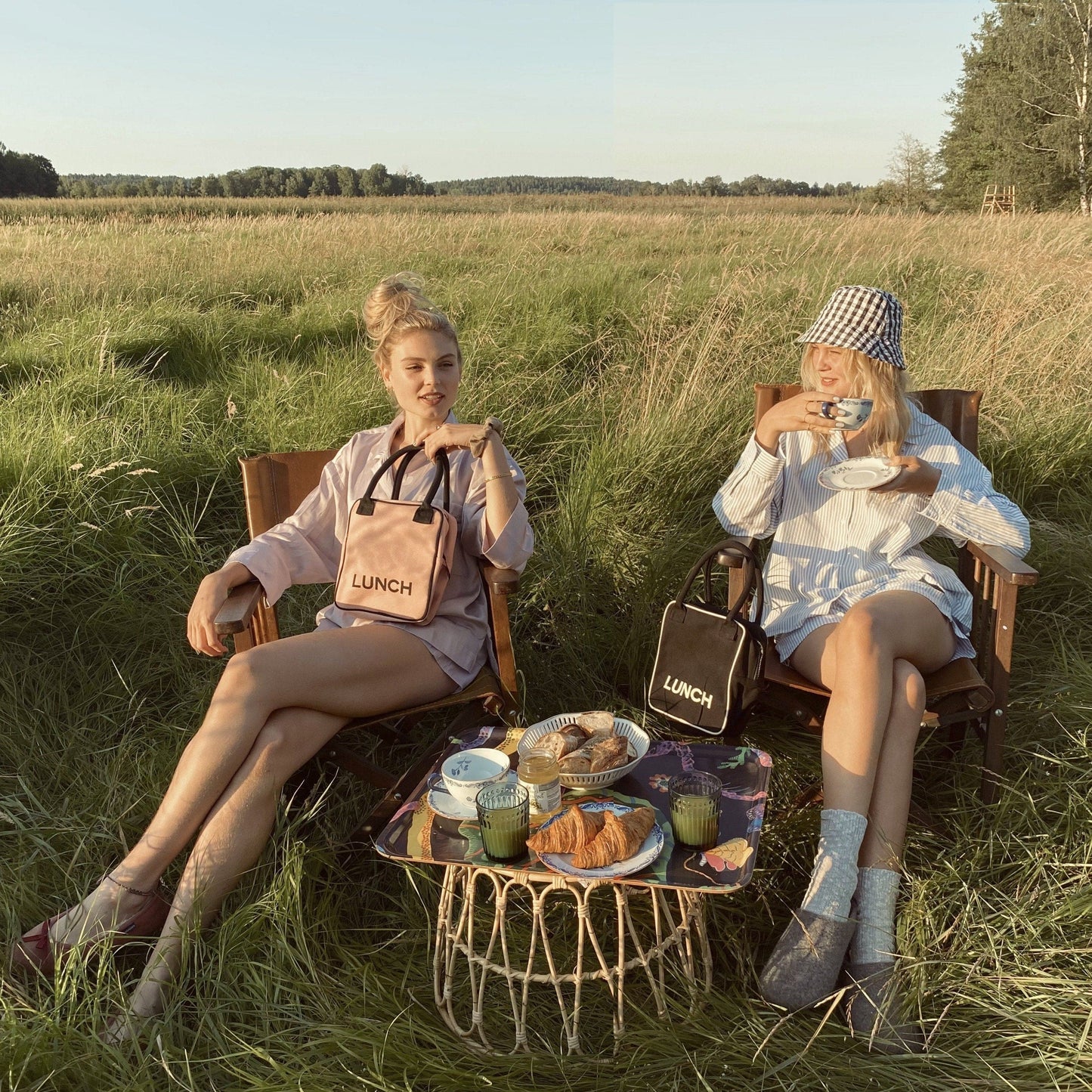 Bag-all Lunch Box Insulated Pink/Blush shown in outdoor picnic setting with snacks and drinks on wicker table, displayed in sunny field with tall grass and forest backdrop