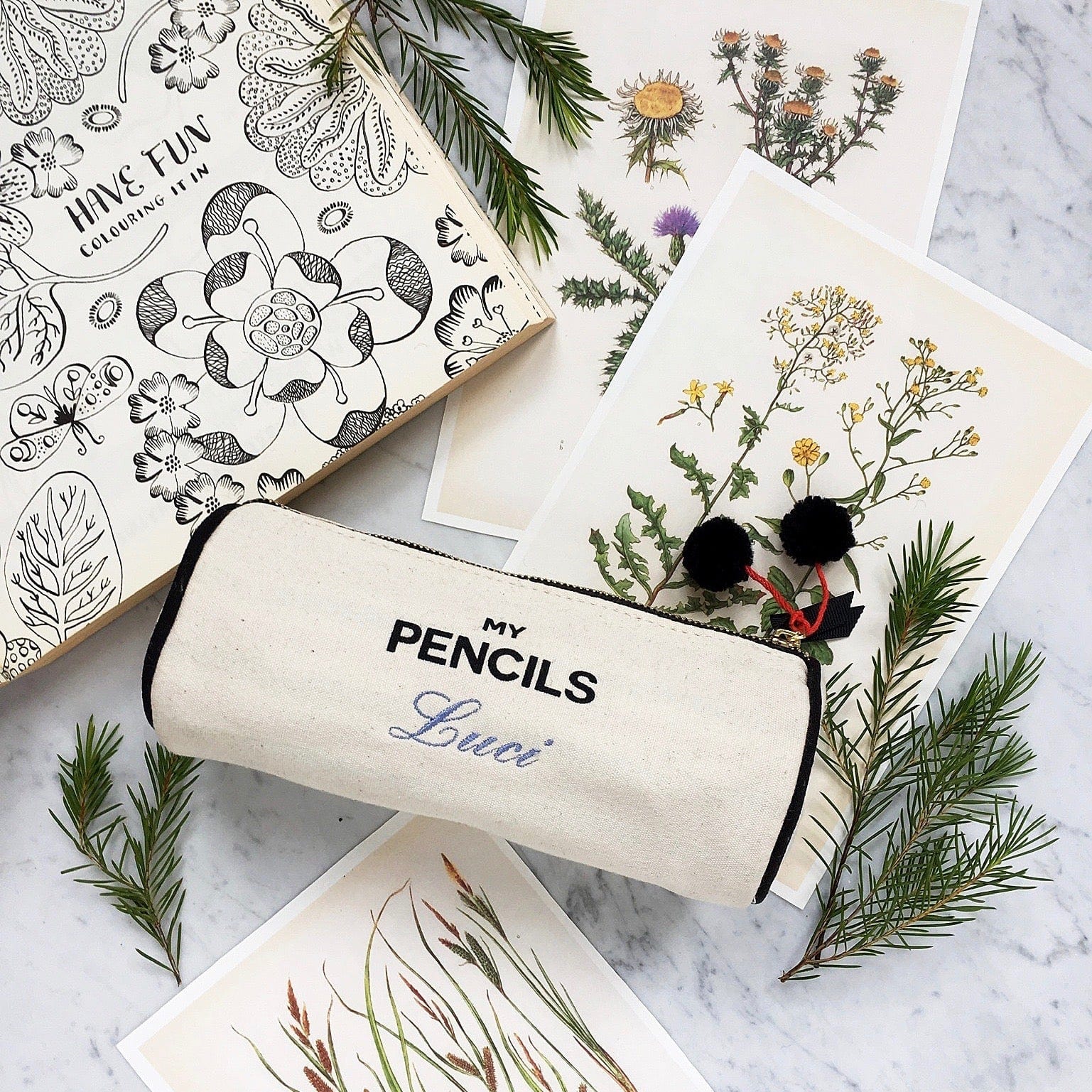 Bag-all cream canvas pencil case with 'My Pencils' text and custom name embroidery, displayed with botanical illustrations and pine sprigs on marble surface